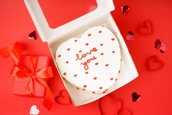 Opened box with heart-shaped bento cake and gift on red background. Valentine's Day celebration