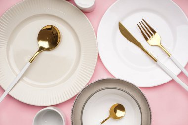 Different clean dishware with golden cutlery on pink background
