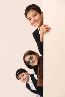 Little friends in stylish school uniform with blank poster near light wall