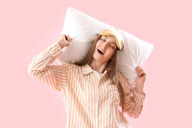 Young woman with sleeping mask and soft pillow on pink background