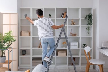 Young man on ladder near bookshelf at home, back view
