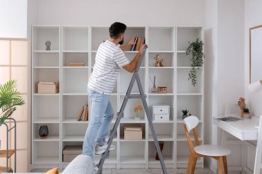 Young man on ladder taking book from shelf at home