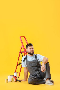 Male worker with ladder and cans of paint sitting on yellow background
