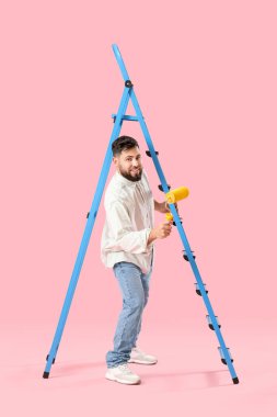 Young man with paint roller and ladder on pink background