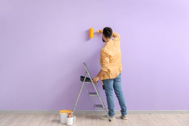 Young man with ladder and roller painting lilac wall, back view