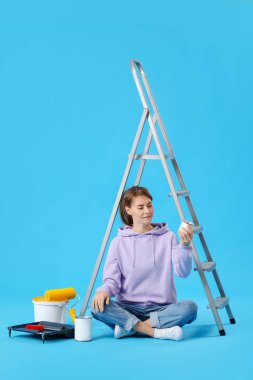 Young woman with paint cans and ladder sitting on blue background