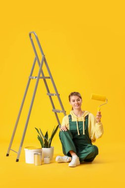 Young woman with roller, ladder, houseplant and paint cans on yellow background