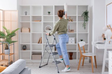 Young woman on stepladder cleaning bookshelf with duster at home