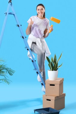 Young woman with paint roller, ladder and moving boxes showing thumb-up on blue background