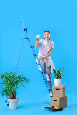 Young woman with paint cans, ladder and moving boxes on blue background