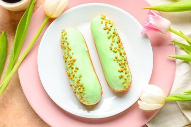 Plate with delicious eclairs and beautiful flowers on light background