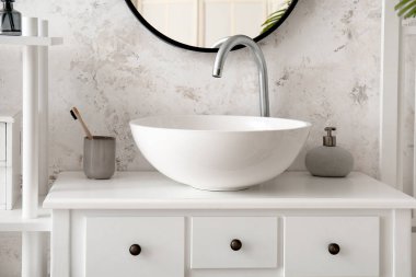 Ceramic sink, holder with toothbrush and soap on table in light bathroom