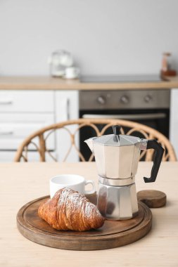 Board with geyser coffee maker and croissant on table in kitchen