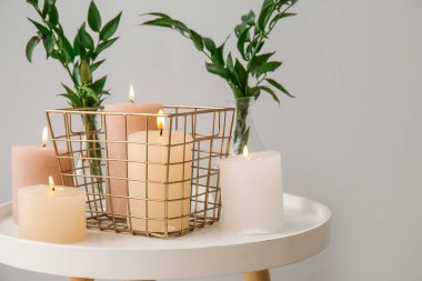 Burning candles and vases with ruscus branches on end table near grey wall