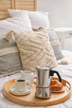 Tray with geyser coffee maker, cup of espresso and delicious croissant on bed