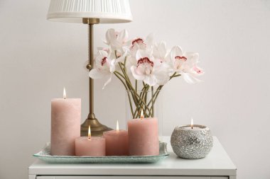 Burning candles, vase with orchid and lamp on bedside table near white wall