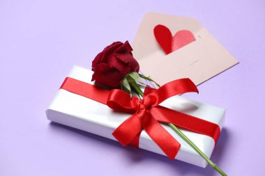 Gift box with beautiful bow, rose flower and envelope on lilac background. Valentine's Day celebration