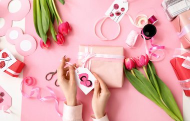 Female hands with paper tag, gift boxes, tulip flowers and packing materials on pink background. International Women's Day celebration