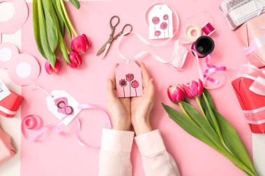 Female hands with paper tag for decorating gift boxes, tulip flowers and packing materials on pink background. International Women's Day celebration
