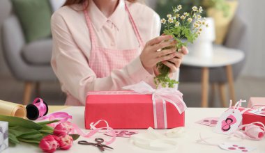 Young woman with chamomile flowers, gift box and packing materials at table. International Women's Day celebration