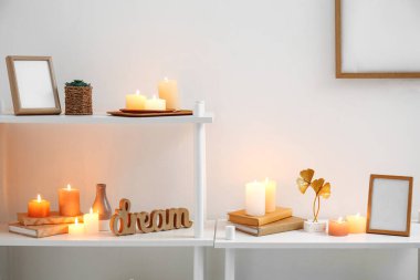 Shelving units with burning candles, books and frames near light wall