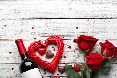 Bottle of wine, chocolate candies, heart and rose flowers on white wooden background. Valentine's Day celebration
