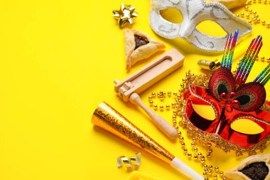 Hamantaschen cookies, carnival masks and noisemakers for Purim holiday on yellow background