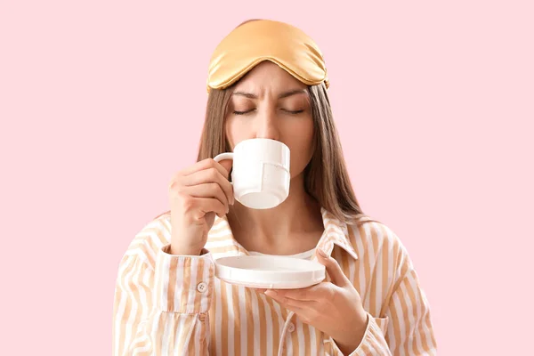 Young woman with sleeping mask drinking coffee on pink background
