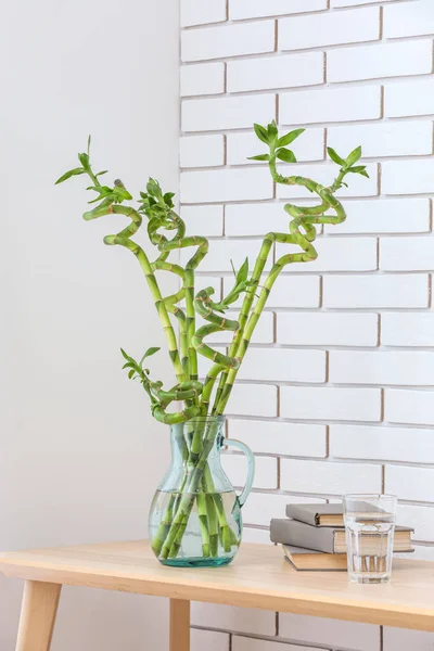 Vase with bamboo branches, books and glass of water on table near light brick wall