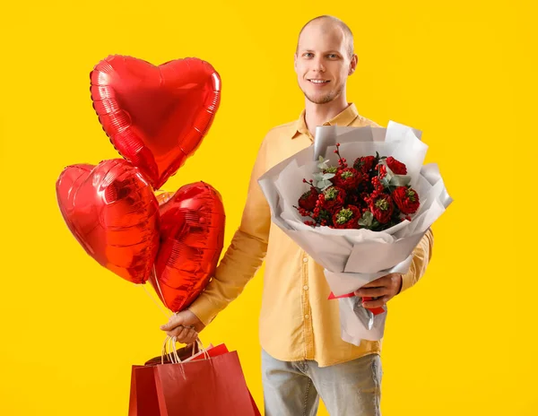 Young man with bouquet of flowers, bags and balloons on yellow background. Valentine\'s Day celebration