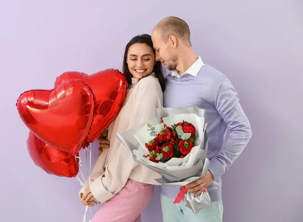 Young couple with bouquet of flowers and balloons on lilac background. Valentine\'s Day celebration