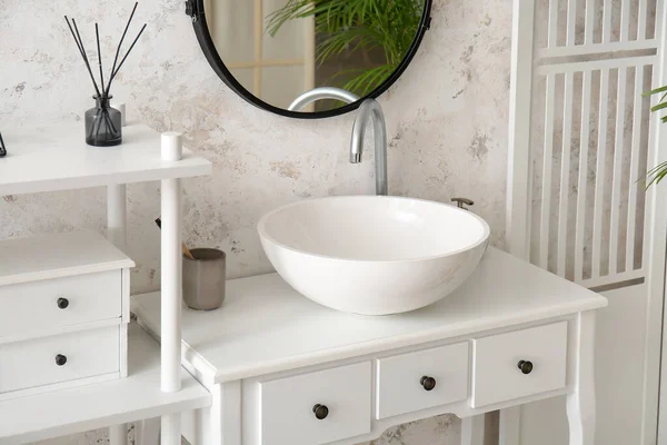 Ceramic sink, holder with toothbrush and soap on table in light bathroom
