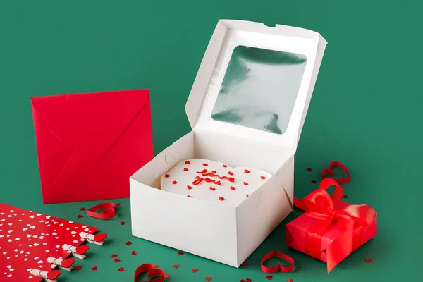 Opened box with tasty bento cake, gift and envelope on green background. Valentine\'s Day celebration