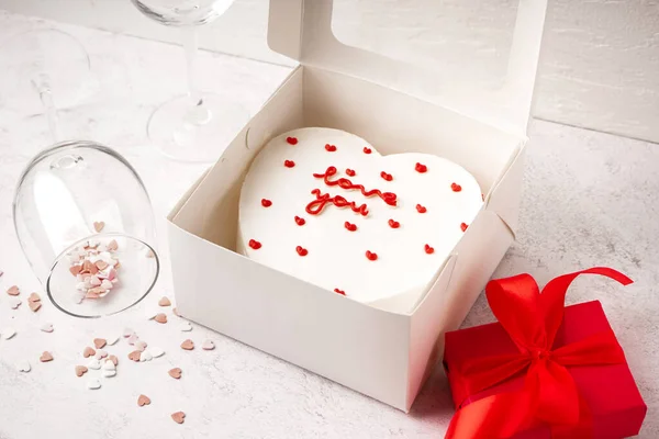 Box with heart-shaped bento cake, glasses and gift on white table. Valentine\'s Day celebration