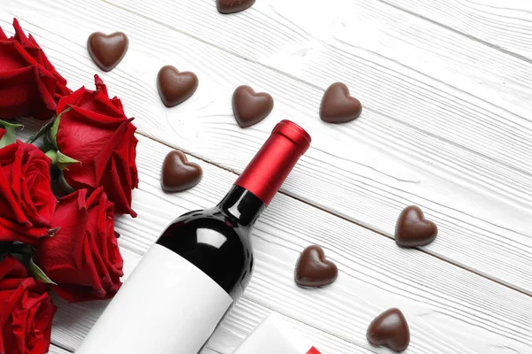 Bottle of wine, chocolate candies and rose flowers on white wooden background. Valentine\'s Day celebration