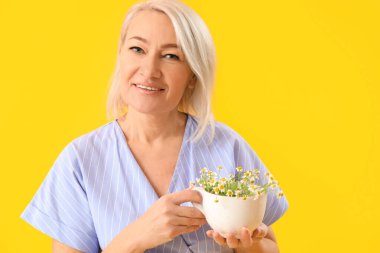 Beautiful mature woman holding cup with chamomiles on yellow background