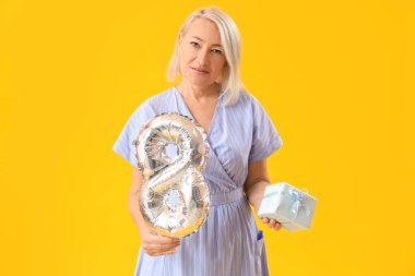 Beautiful mature woman with balloon in shape of figure 8 and gift on yellow background. International Women's Day