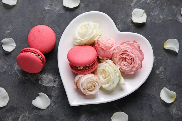 Heart-shaped plate with tasty macaroons and rose flowers on black background. Valentine's Day celebration