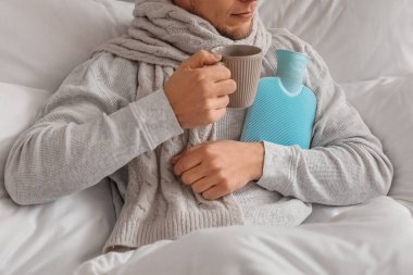 Ill man with cup of tea and hot water bottle in bedroom, closeup