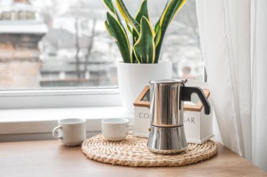 Wicker coaster with geyser coffee maker and cups on table near window