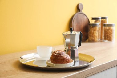 Tray with geyser coffee maker, delicious bun and cup on kitchen counter near yellow wall