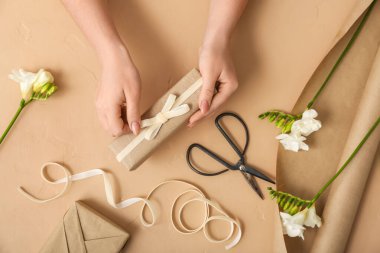 Female hands with gift box, wrapping paper, ribbon, scissors and freesia flowers on color background. Women's Day celebration