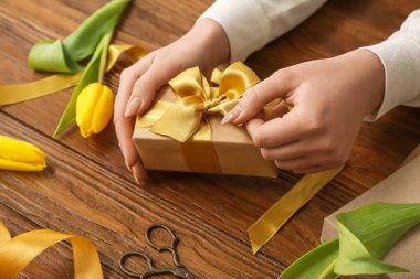Female hands with beautiful gift box and tulip flowers on wooden background, closeup. Women's Day celebration