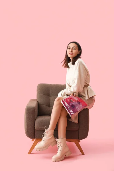 Young woman with magazine sitting in soft armchair on pink background