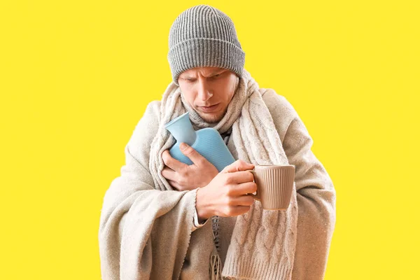 Frozen young man with hot water bottle and plaid on yellow background