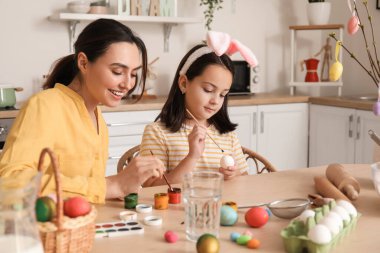 Little girl and her mother painting Easter eggs at table in kitchen