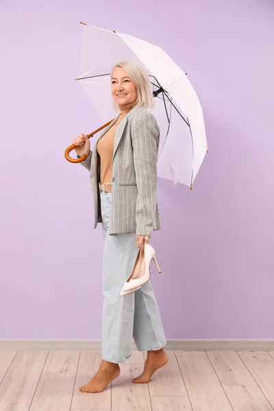 Barefoot mature woman with umbrella on lilac background