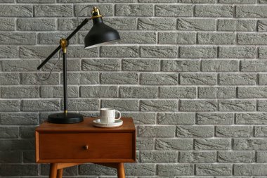 Wooden table with drawer, lamp and cup of coffee near grey brick wall