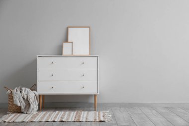 White chest of drawers with blank frames and basket near grey wall