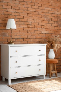 Chest of drawers with lamp, stool and vase near brick wall